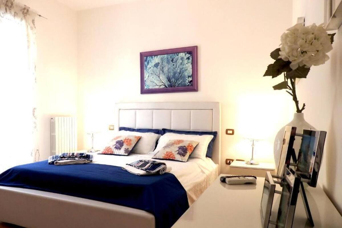 A neatly made bed with a blue bedspread, a framed picture on the wall, and a vase with white flowers on a bedside table.
