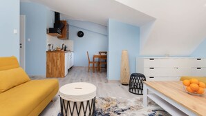 A modern living room with a yellow sofa, a wooden dining table with oranges, and a kitchen area with a wooden counter and a TV. - Comfortable Studio in Puck | Parking and 400 m to the Beach (Puck)