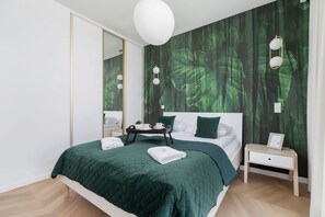 Une chambre moderne avec un mur aux tons verts, un lit recouvert d’une courtepointe verte matelassée, une table de chevet avec une lampe et une petite table de nuit avec un cadre photo.