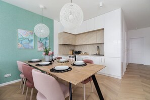 A modern dining area with a wooden table set for four, surrounded by pink chairs. There are two pendant lights above the table, and the room has white cabinets and a green accent wall.