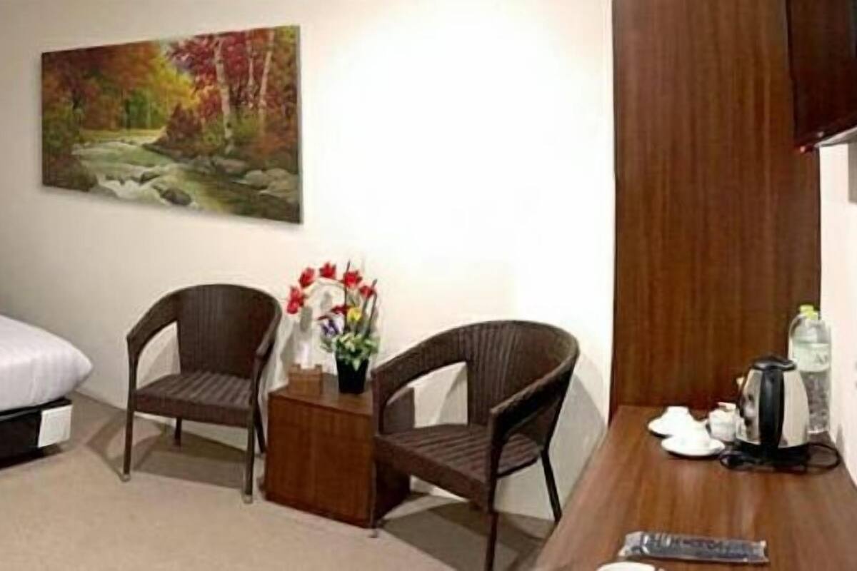 A hotel room with a bed, two wicker chairs, a small wooden table with a vase of flowers, and a desk with a kettle and a bottle.