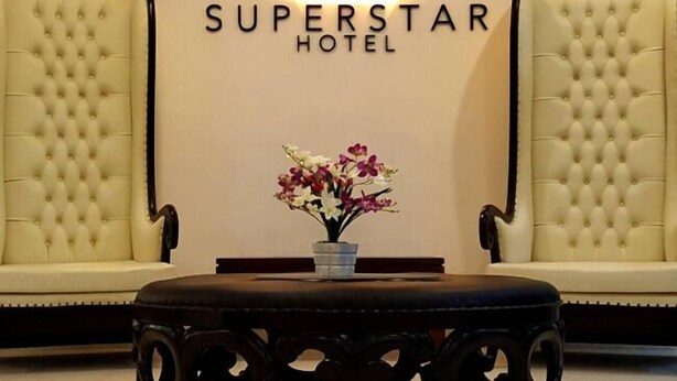 Een hotel lobby met twee comfortabele fauteuils, een bijzettafeltje met een bloemenvaas, en een neonbord waarop 'SUPERSTAR HOTEL' staat.
