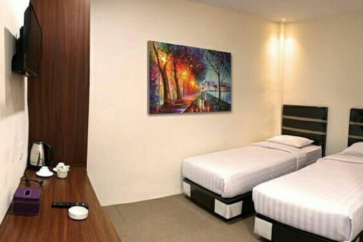 A hotel room with two single beds, a painting on the wall, a desk with a kettle, and a small table with a vase.