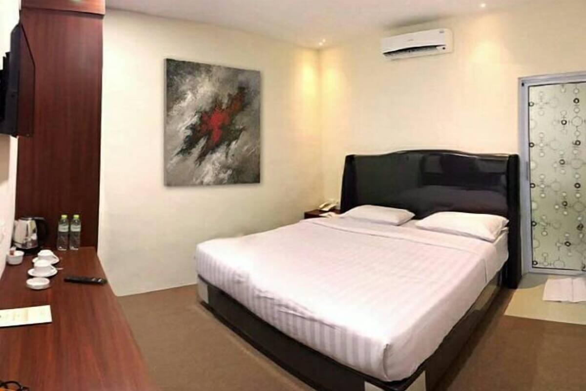 A hotel room with a large bed, a desk with a chair, a painting on the wall, and an air conditioning unit.