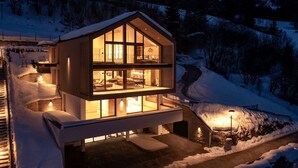 Moderni, kaksikerroksinen puinen mökki, jossa on suuret ikkunat, parveke ja lumipeitteinen maisema.