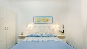 Un dormitorio con cama, paredes blancas, un cuadro, dos lámparas de noche y una mesita de noche con una planta.