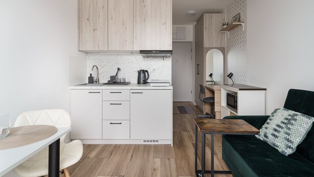 Cocina compacta con armarios blancos, encimera de madera y horno integrado. Hay un comedor con una silla blanca y una mesa, y un sofá verde con un cojín estampado.