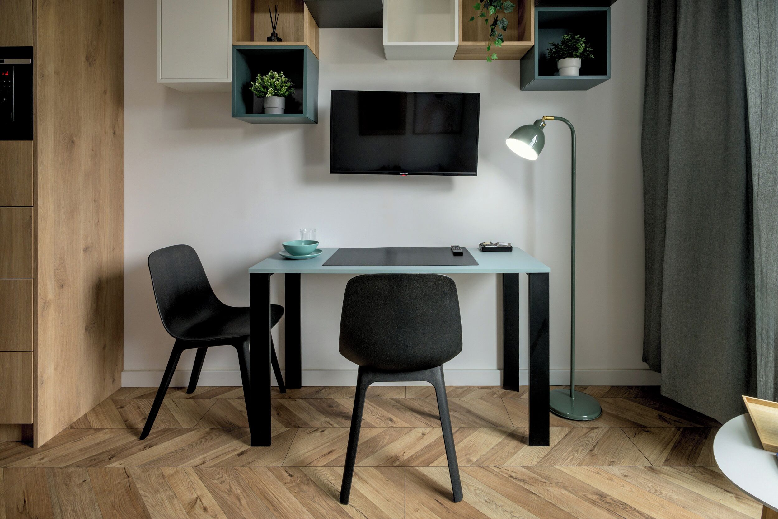 A modern room with a wooden herringbone floor, a desk with two black chairs, a flat-screen TV mounted on the wall, and a floor lamp.