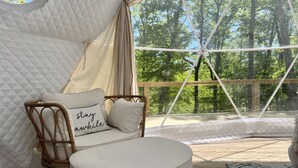 A cozy, well-lit tent with a cushioned seating area, a wooden chair, and a view of a forest through a large window. - Glamping Dome · Hot Tub, Fire Pit & Forest Views (Simpson)