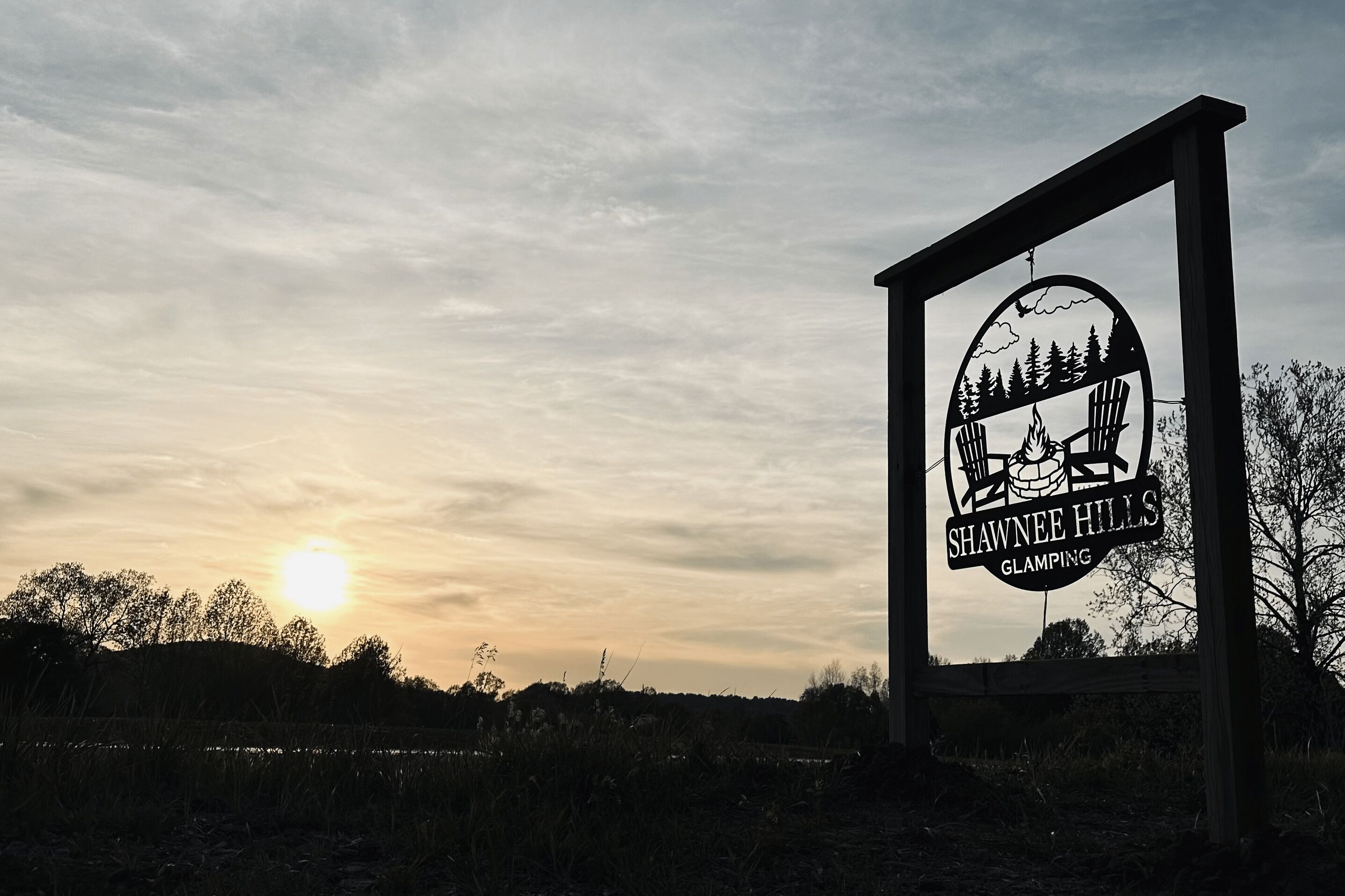 A sign with the text "SHAWNEE HILLS GLAMPING" against a sunset sky.