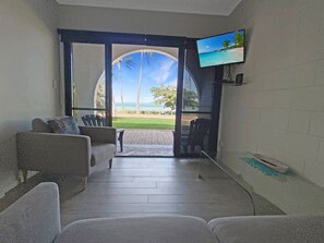 A room with a TV, a sofa, and a view of a beach. - Beachside Palms Unit 3 (Nelly Bay)
