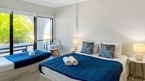 Habitació d'hotel amb un llit gran, un llit més petit, dues nits abelles amb làmpades, una finestra amb vista a palmeres i un balcó.