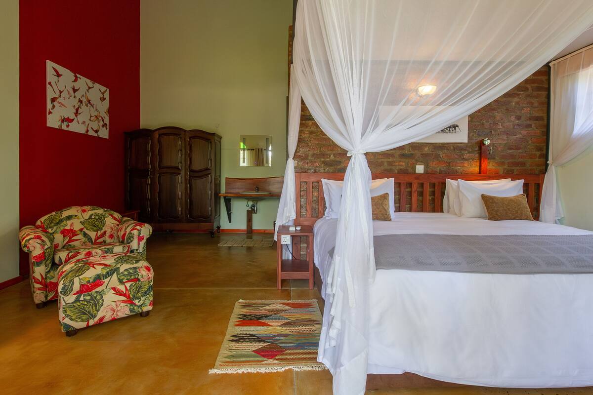 Un dormitorio con una cama con dosel, una mesita de noche de madera, un sillón tapizado con flores y una pared roja con un cuadro de un pájaro.