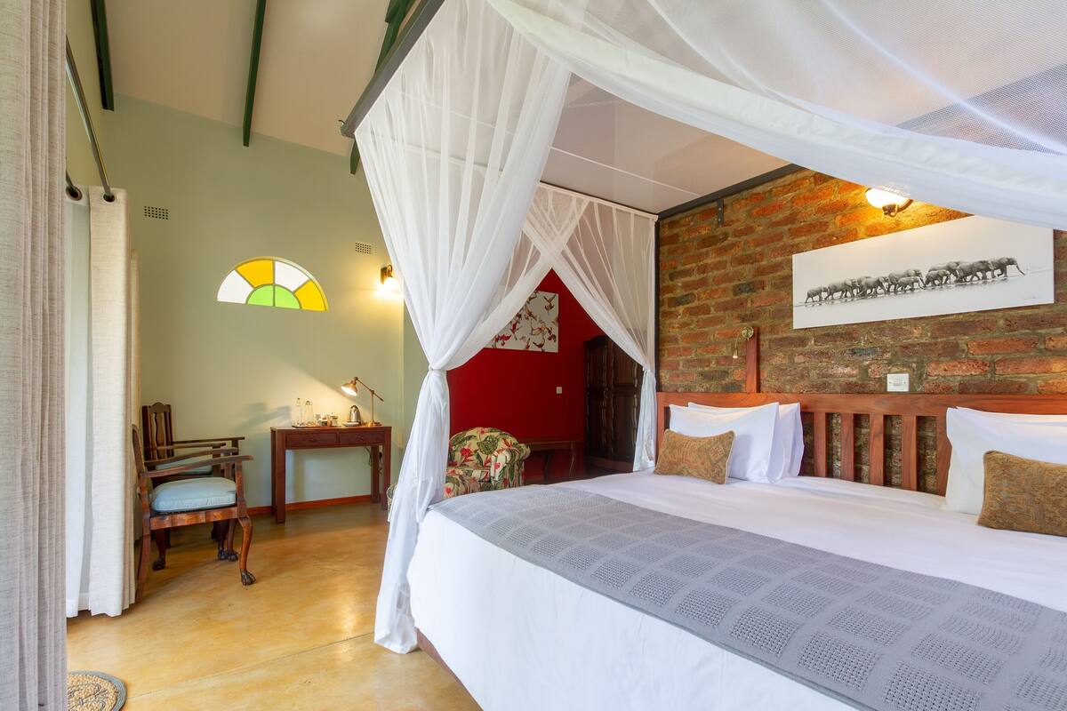 Una habitación con cama, una silla, un escritorio, un cuadro en la pared y una ventana con un diseño de vitral.
