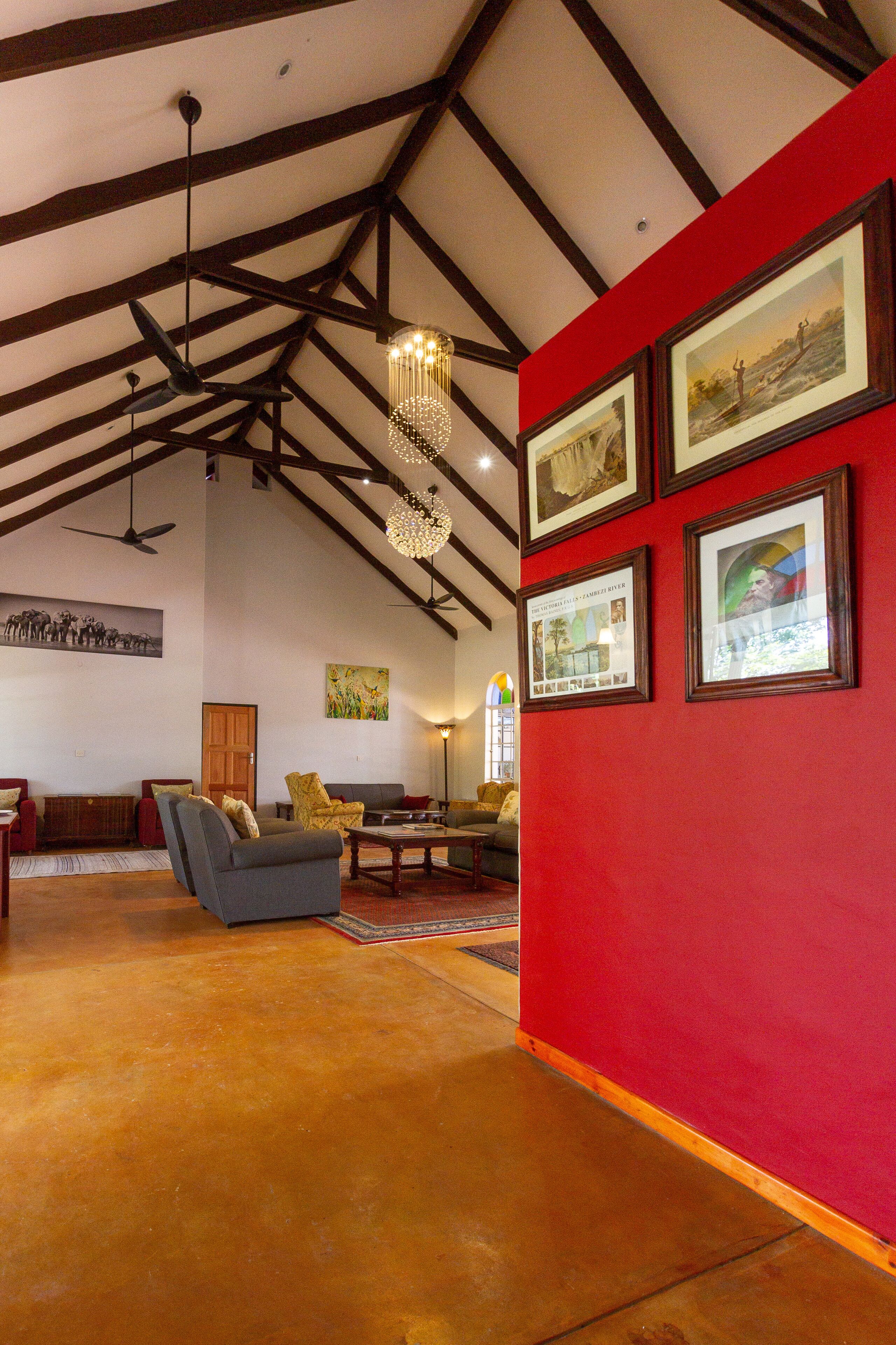 A spacious living area with a high ceiling, exposed beams, and a red accent wall. There are framed pictures on the wall and a chandelier hanging from the ceiling.