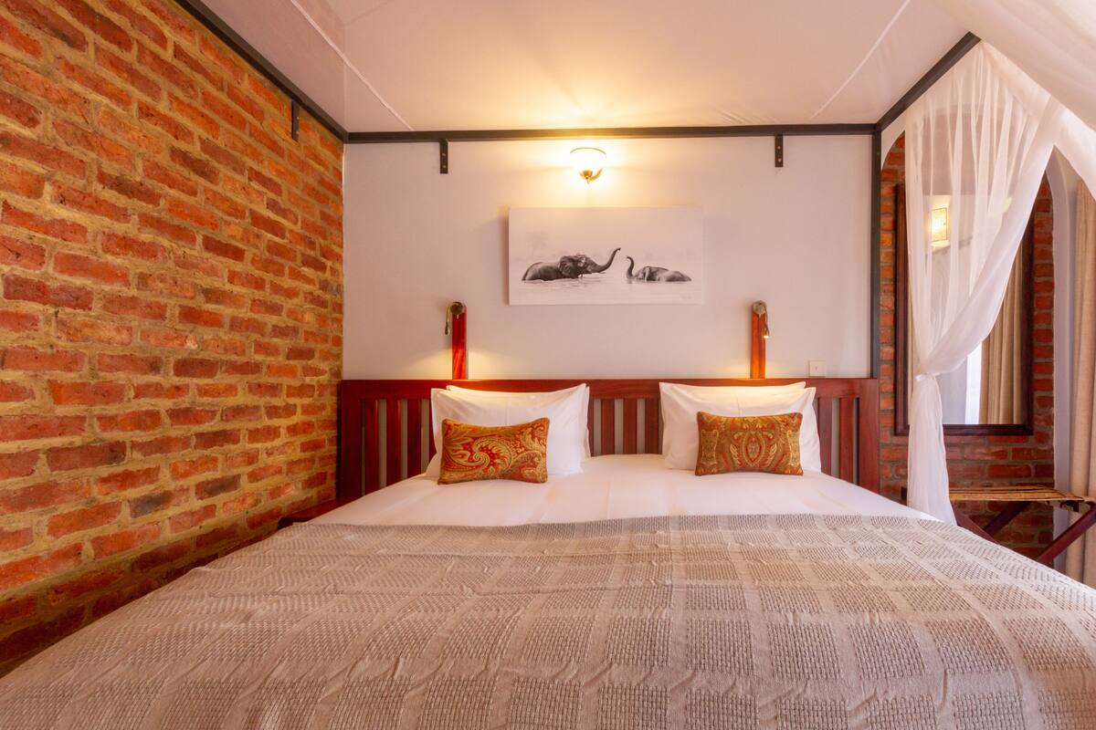 Habitación de hotel con pared de ladrillo, cama con ropa blanca y almohadas estampadas, una obra de arte en la pared con elefantes y una cama con dosel y cortinas traslúcidas.
