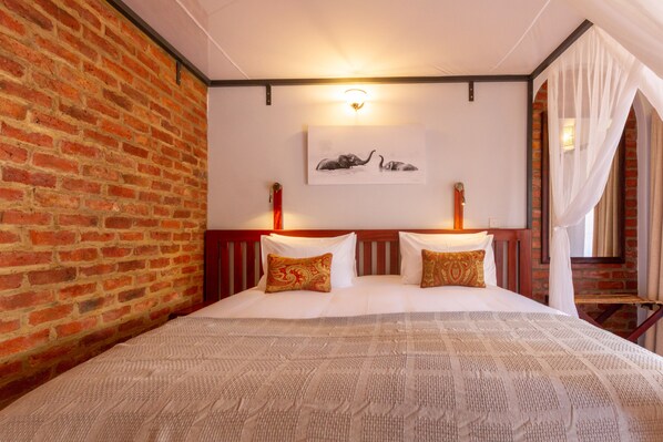 Habitación de hotel con pared de ladrillo, cama con ropa blanca y almohadas estampadas, una obra de arte en la pared con elefantes y una cama con dosel y cortinas traslúcidas.