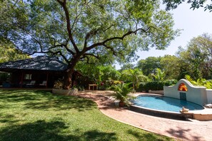 Un patio trasero con piscina, un espacio de estar techado y un gran árbol que da sombra.