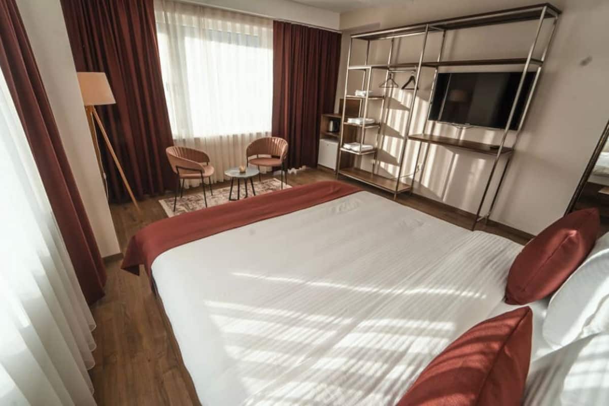 A hotel room with a large bed, a TV mounted on a wall, a seating area with chairs, and a wardrobe with shelves.