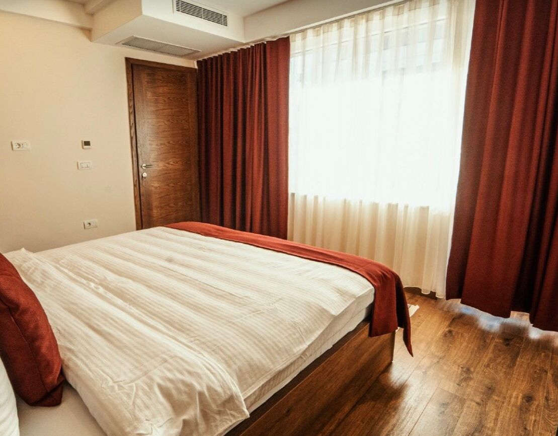 Un dormitorio con cama de madera, cortinas rojas y una ventana con cortinas traslúcidas.