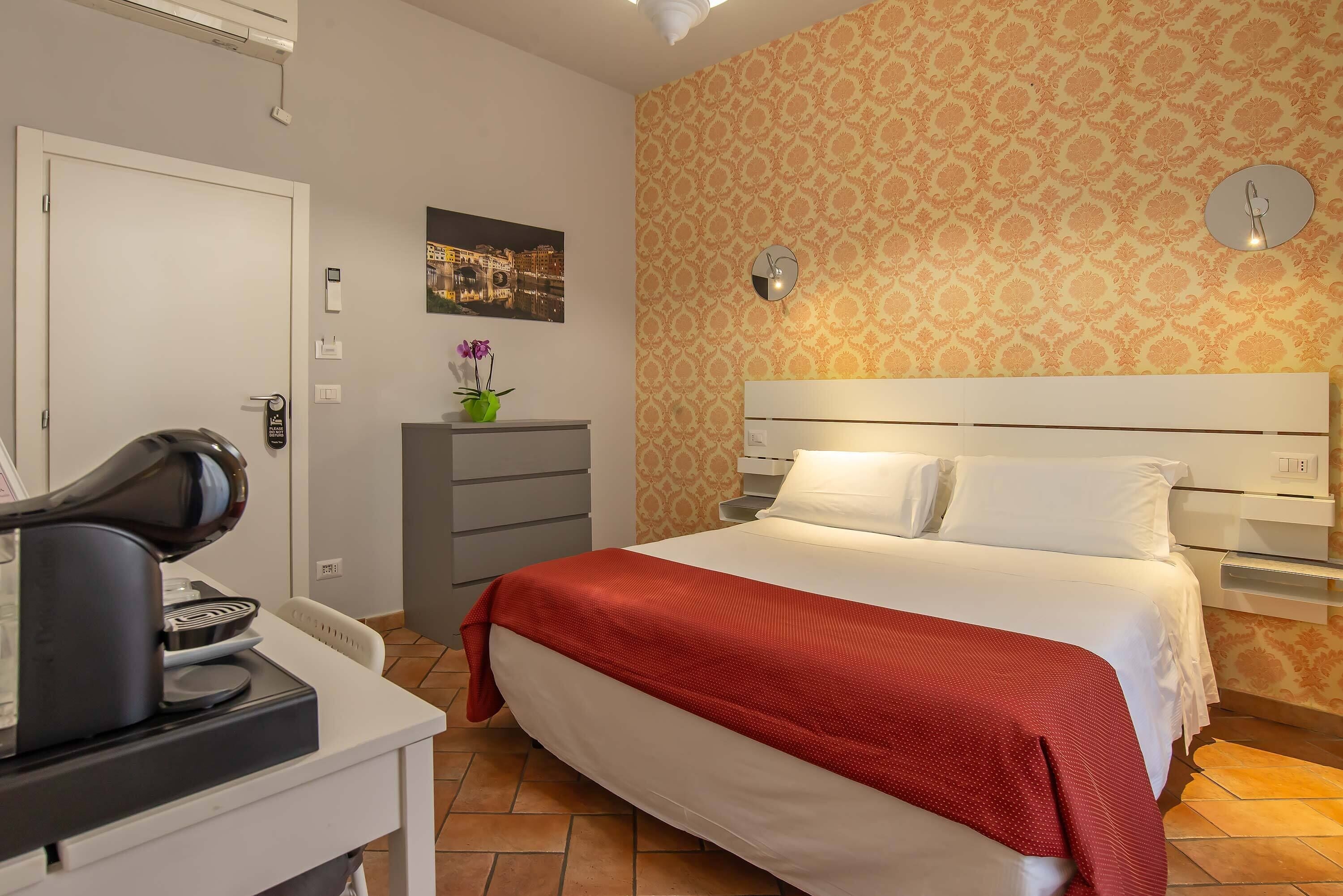 Hotellihuone, jossa on sänky, työpöytä, kahvinkeitin, seinäkello ja kuviollinen tapetti.