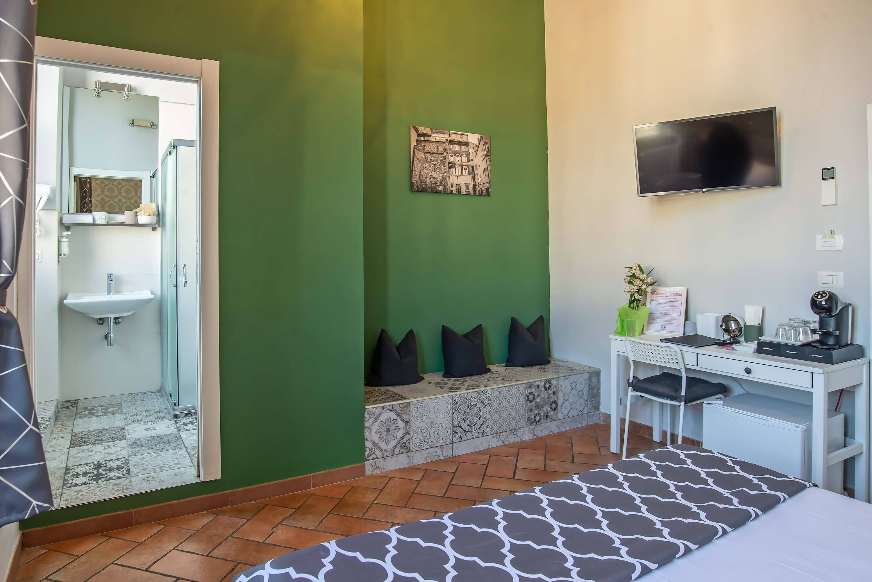 Une chambre d’hôtel avec un mur vert, un lit recouvert d’une couvre-lit à motifs, un bureau avec une chaise, un téléviseur à écran plat, un lavabo et un banc avec des coussins.