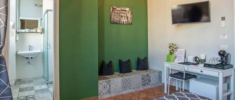 Une chambre d’hôtel avec un mur vert, un lit recouvert d’une couvre-lit à motifs, un bureau avec une chaise, un téléviseur à écran plat, un lavabo et un banc avec des coussins.