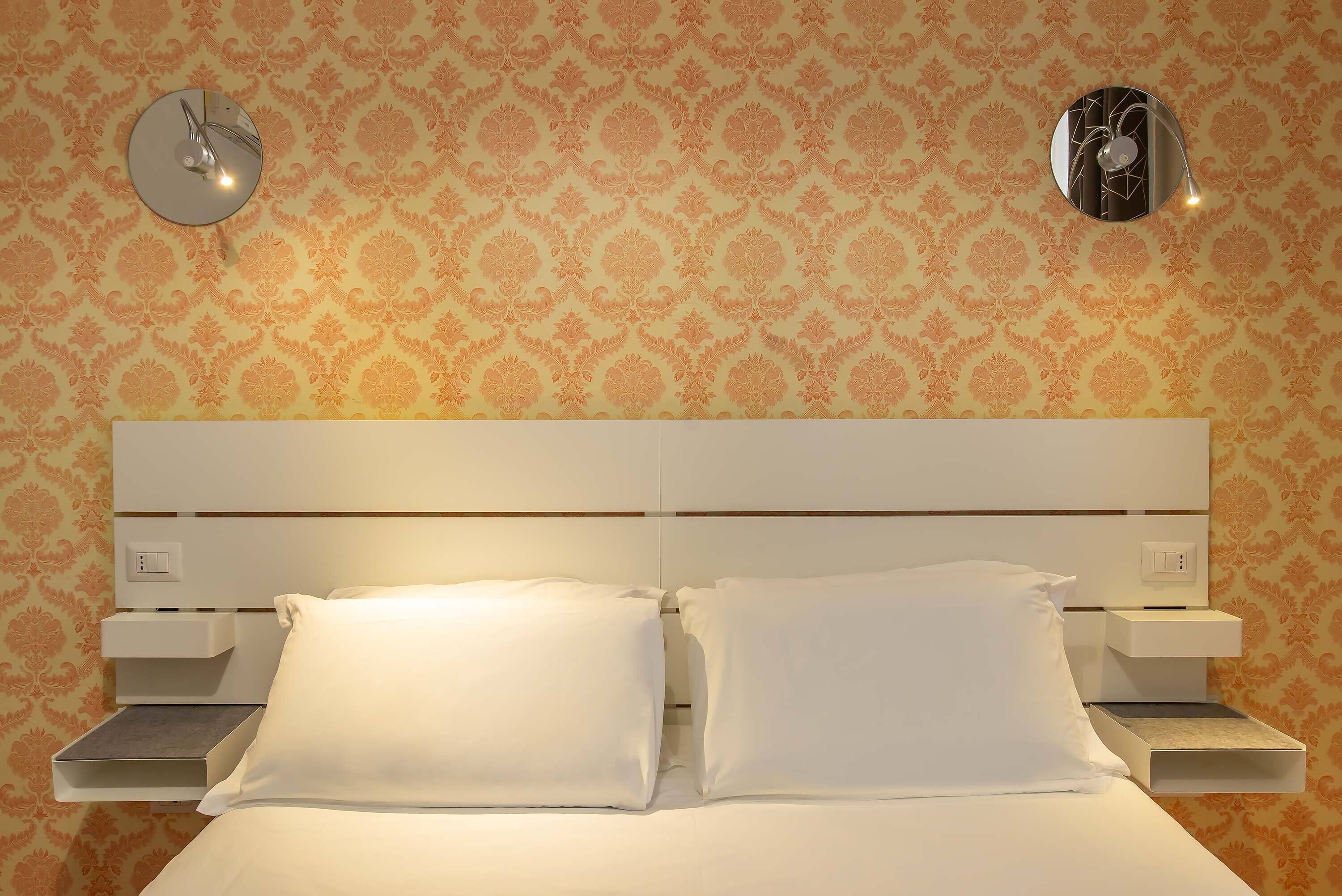 Una habitación de hotel con papel tapiz estampado, una cama con sábanas blancas y dos mesitas de noche.