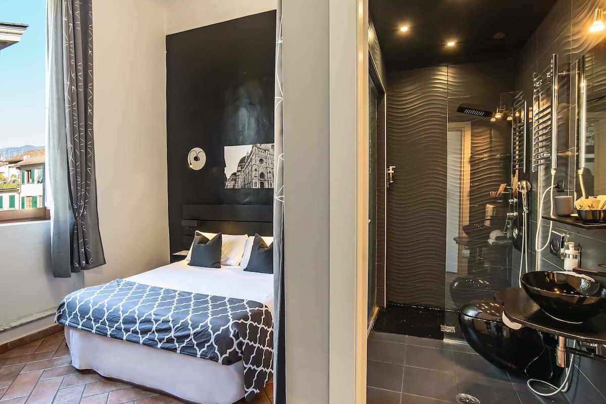 Une chambre d’hôtel moderne dotée d’un grand lit, d’une couvre-lit à motifs noir et blanc, d’une œuvre d’art représentant un paysage urbain accrochée au mur, et d’une salle de bain contemporaine avec un lavabo noir et des murs recouverts de miroirs.