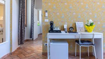 Habitación de hotel compacta con piso de baldosas, un escritorio con silla, un refrigerador pequeño, una cafetera, un florero con flores y un certificado enmarcado sobre el escritorio.