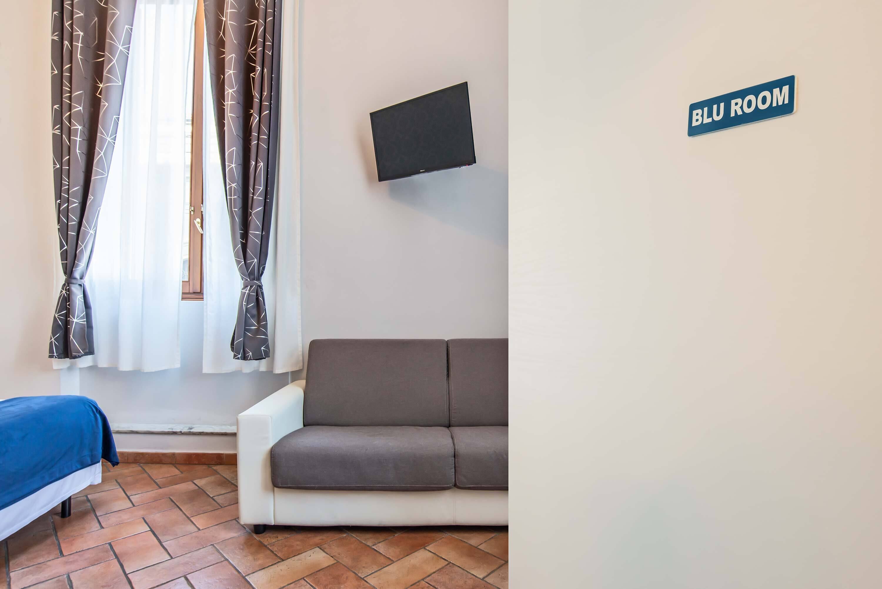 A room with a bed, a sofa, a TV mounted on the wall, and a sign indicating 'BLU ROOM'.