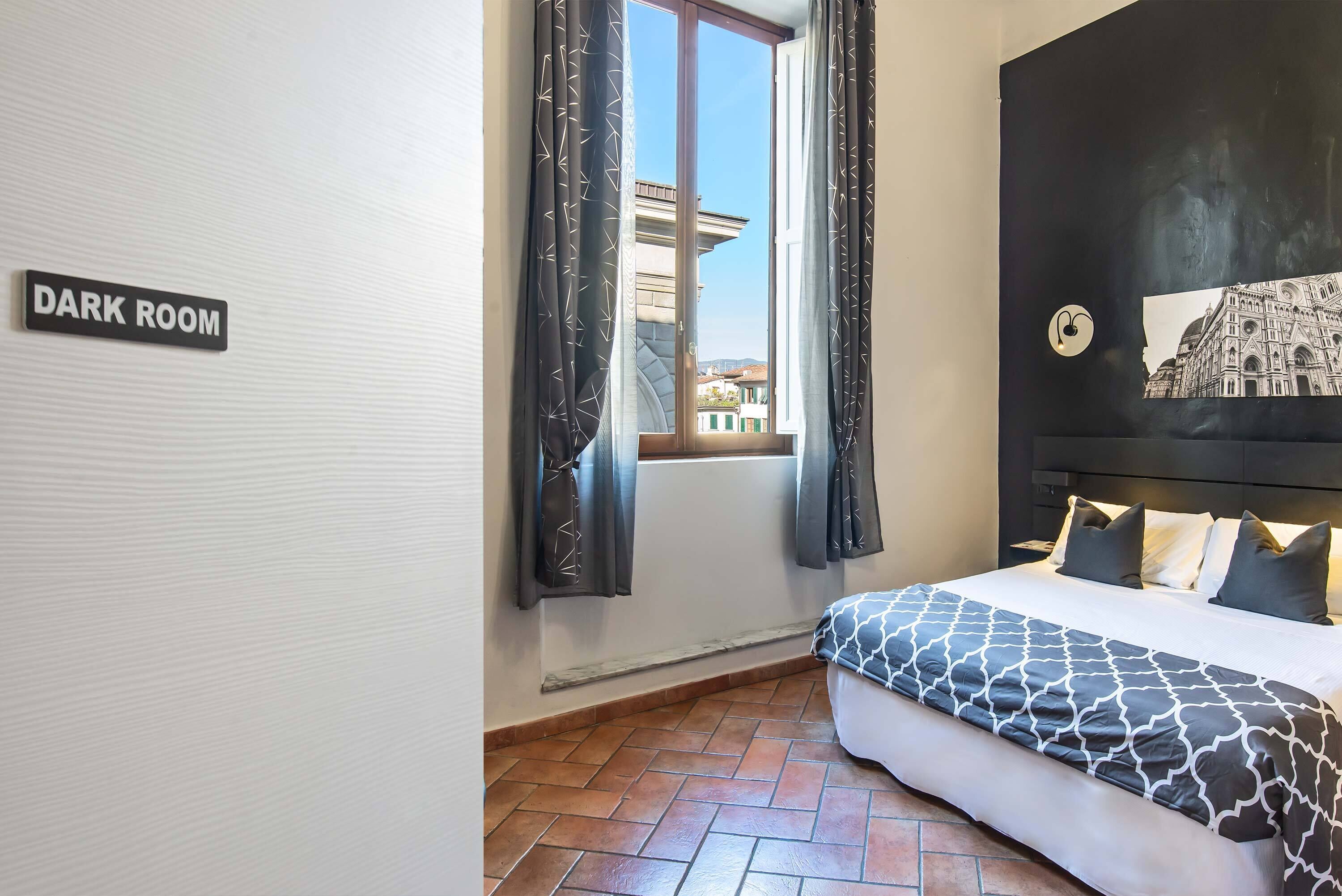 Una habitación oscura con una cama, una ventana con cortinas y vista a la ciudad.