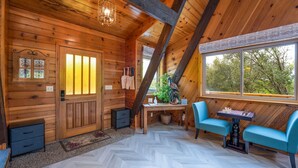 Interior de una cabaña de madera con piso de espiga, una puerta de madera con ventana, una mesa con sillas y un ventanal con vistas a árboles.