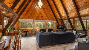 Un loft espacioso con techo alto, vigas de madera, un sofá grande, una mesa de comedor con sillas y grandes ventanales que ofrecen vistas al exterior.