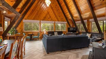 A spacious loft with a high ceiling, wooden beams, a large sofa, a dining table with chairs, and large windows offering a view of the outdoors.