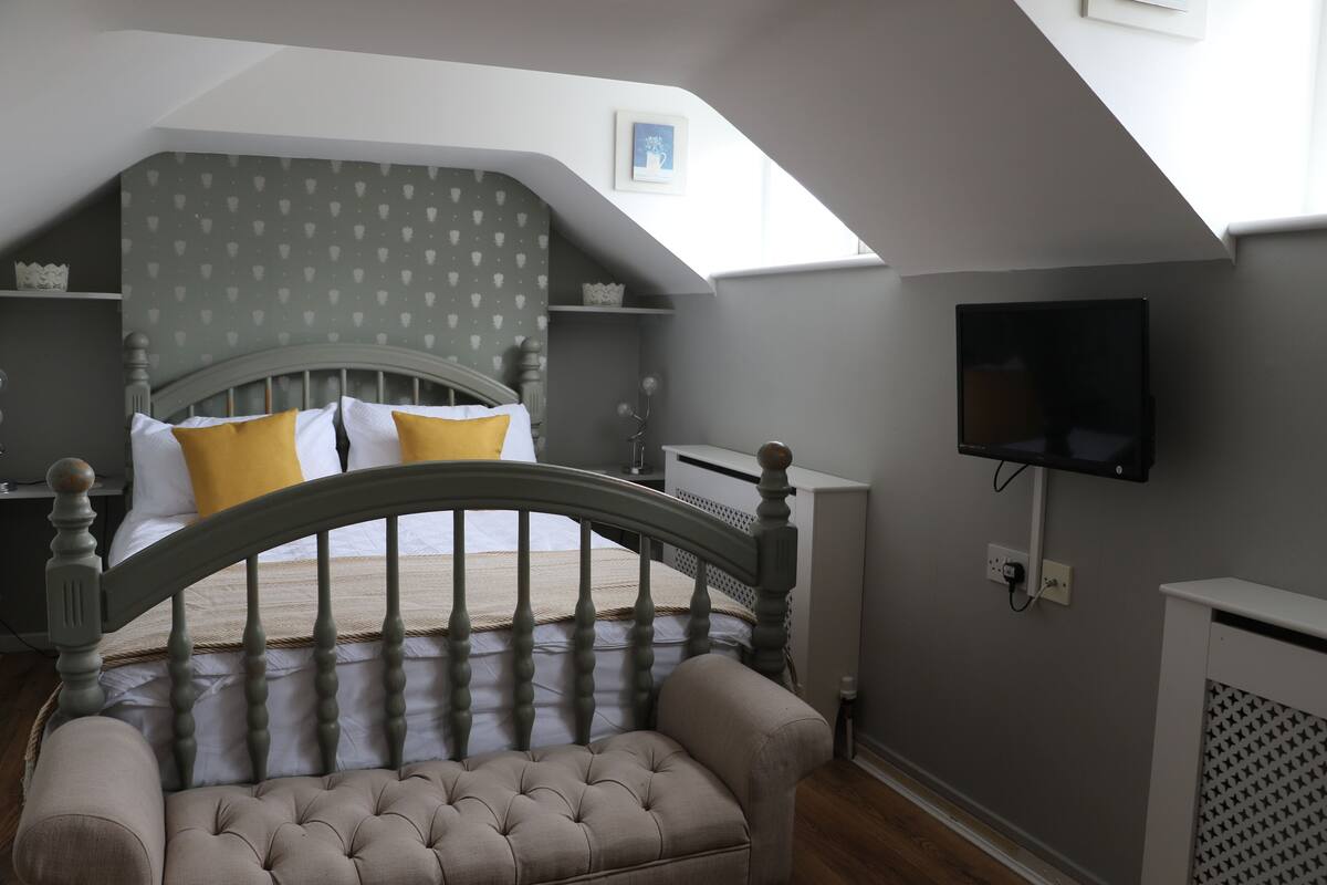 A bedroom with a sloped ceiling, a bed with a wooden headboard, a television mounted on the wall, and a radiator.