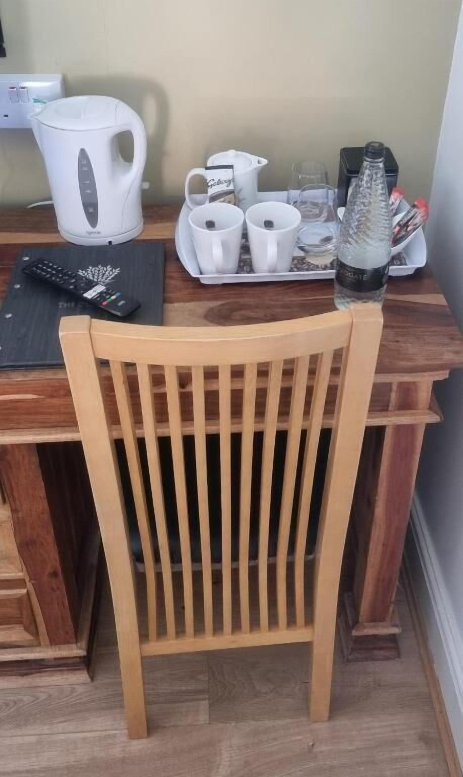 A wooden dining table with a white kettle, two white mugs, a glass pitcher, a bottle of water, and a remote control.
