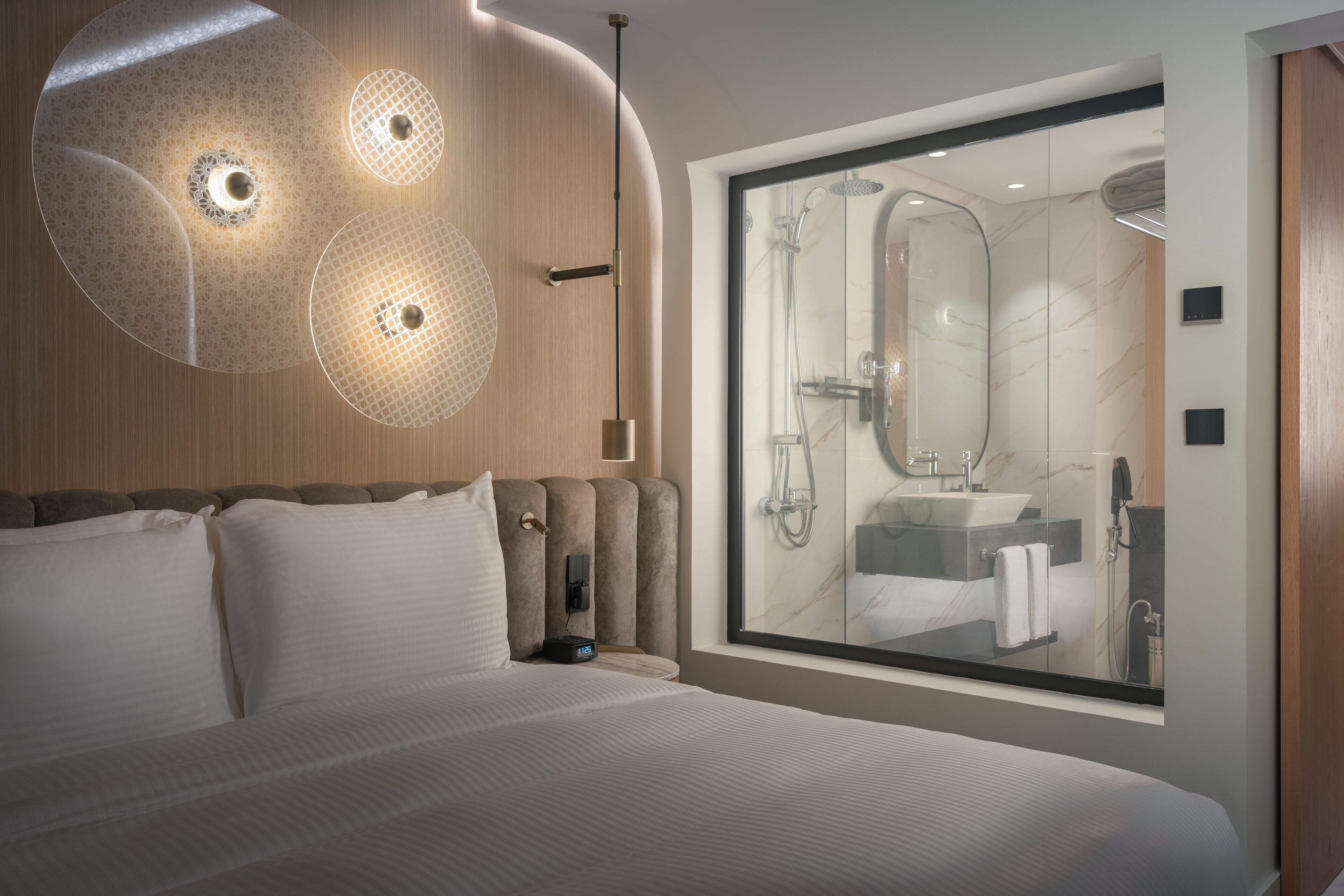 A modern hotel room with a large bed, a glass shower enclosure, and a wall-mounted light fixture.