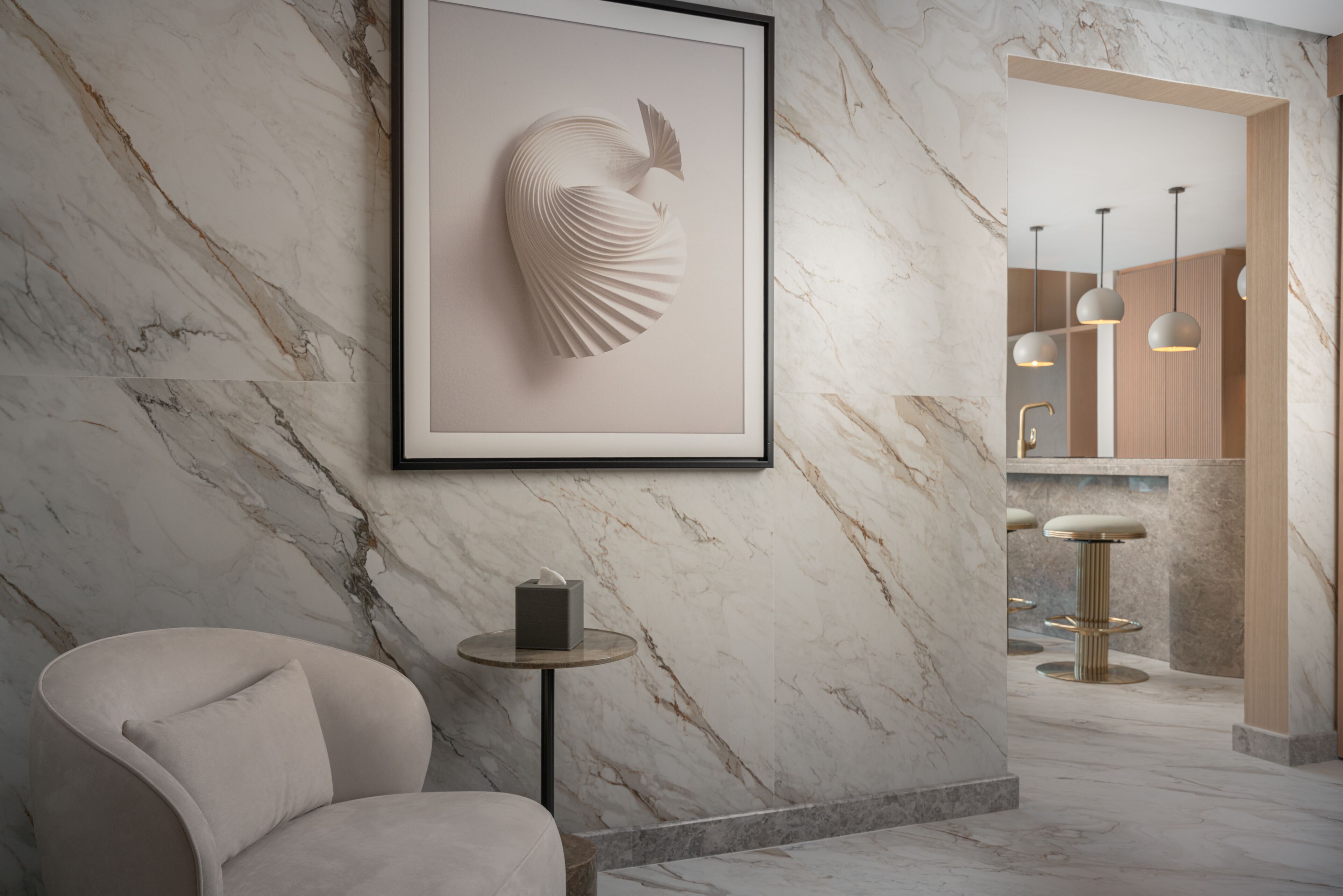 A modern bathroom with a marble wall, a framed artwork, a round table with a tissue box, and a bar area with stools and pendant lights.
