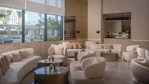 A modern lounge area with a large window offering a view of a pool and palm trees, furnished with sofas and round tables. - Joudyan Jeddah Red Sea Mall (Jeddah)