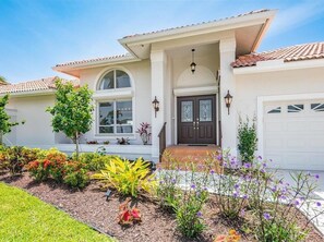 A two-story house with a front porch, a garage, and a well-maintained garden with various plants and flowers. - Waterfront 3BR Marco Island home with heated pool and WiFi. (Marco Island)
