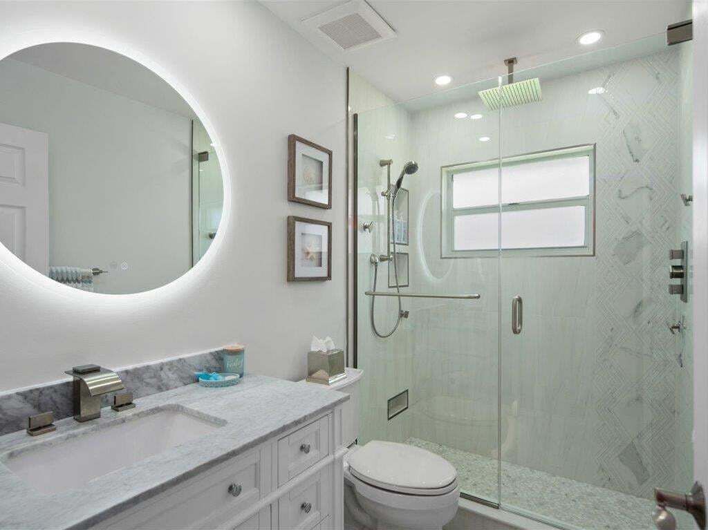 A modern bathroom with a glass-enclosed shower, a round mirror, and a marble vanity.