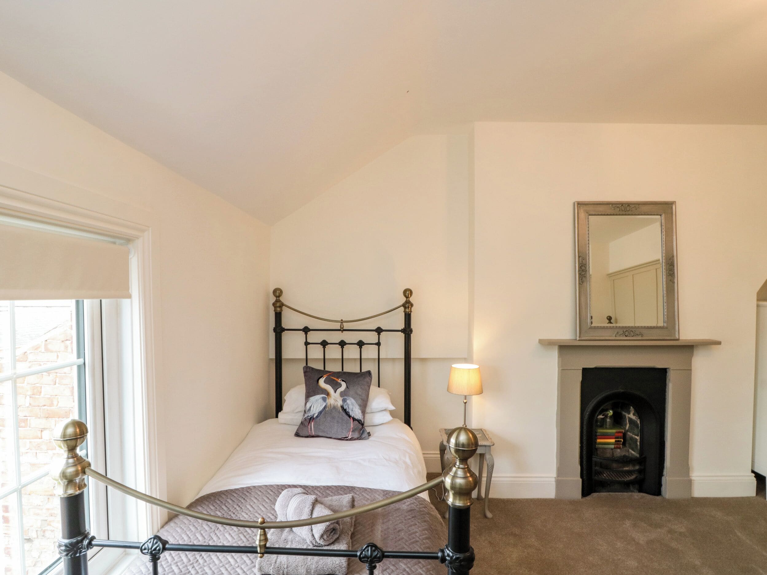 A neatly arranged bedroom with a bed, a bedside lamp, a fireplace, and a window with a view of a brick wall.