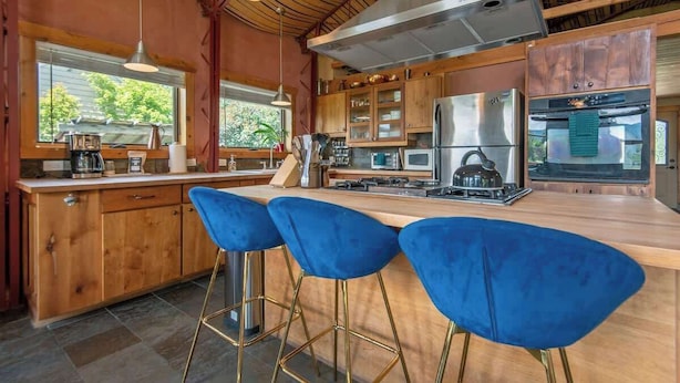 A modern kitchen with wooden cabinets, stainless steel appliances, and a central island with blue bar stools. - Teton Homestead: Cozy Cabin in Downtown Victor (Victor)