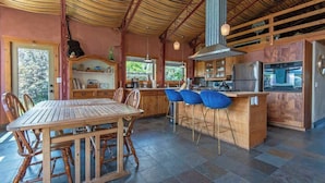A spacious kitchen with a wooden dining table and bar stools, large windows, and a high ceiling with exposed beams. - Teton Homestead: Cozy Cabin in Downtown Victor (Victor)