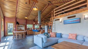 A spacious living area with a large sofa, a wooden coffee table, and a dining table with chairs. There is a kitchen area with a refrigerator and a sink. A ladder is propped against the wall, and a painting is visible on the wall. - Teton Homestead: Cozy Cabin in Downtown Victor (Victor)