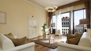 Amplia sala de estar con una mesa de centro de vidrio, dos sofás blancos, piso de madera y vistas a un edificio histórico a través de amplios ventanales.