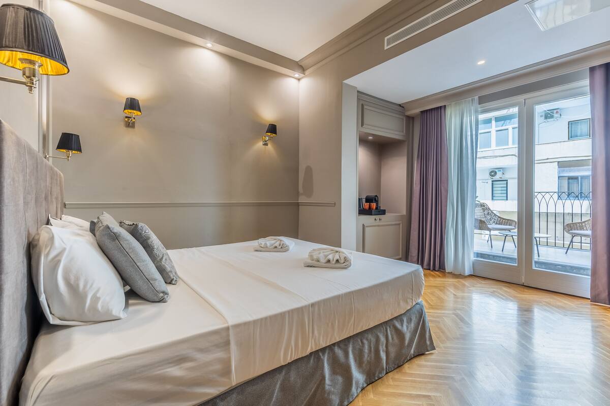 Una habitación de hotel con una cama grande, dos lámparas de pared, una mesita con cafetera y un balcón con mobiliario exterior.