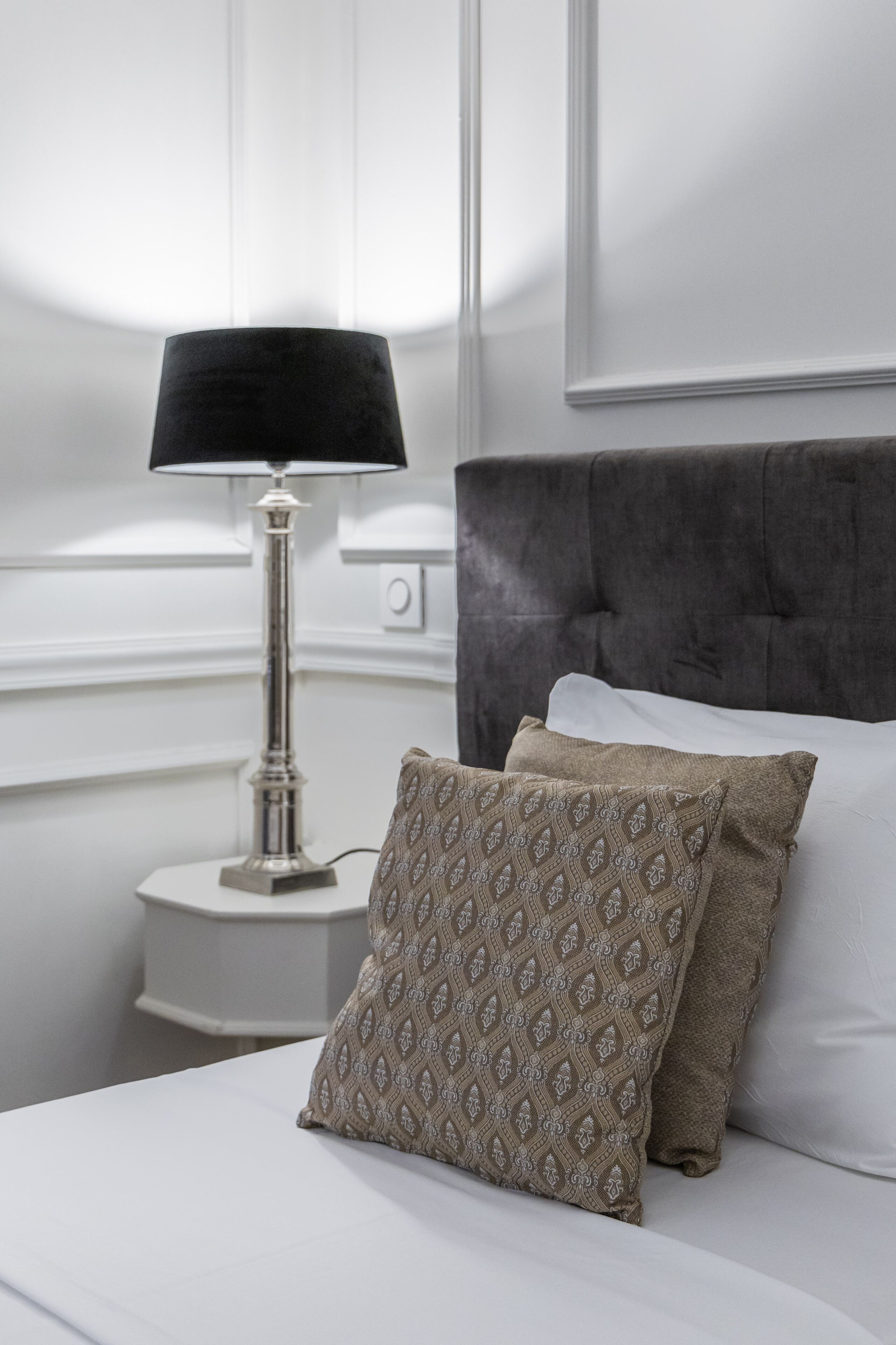 A neatly made bed with a patterned pillow, a bedside lamp, and a dark upholstered headboard.