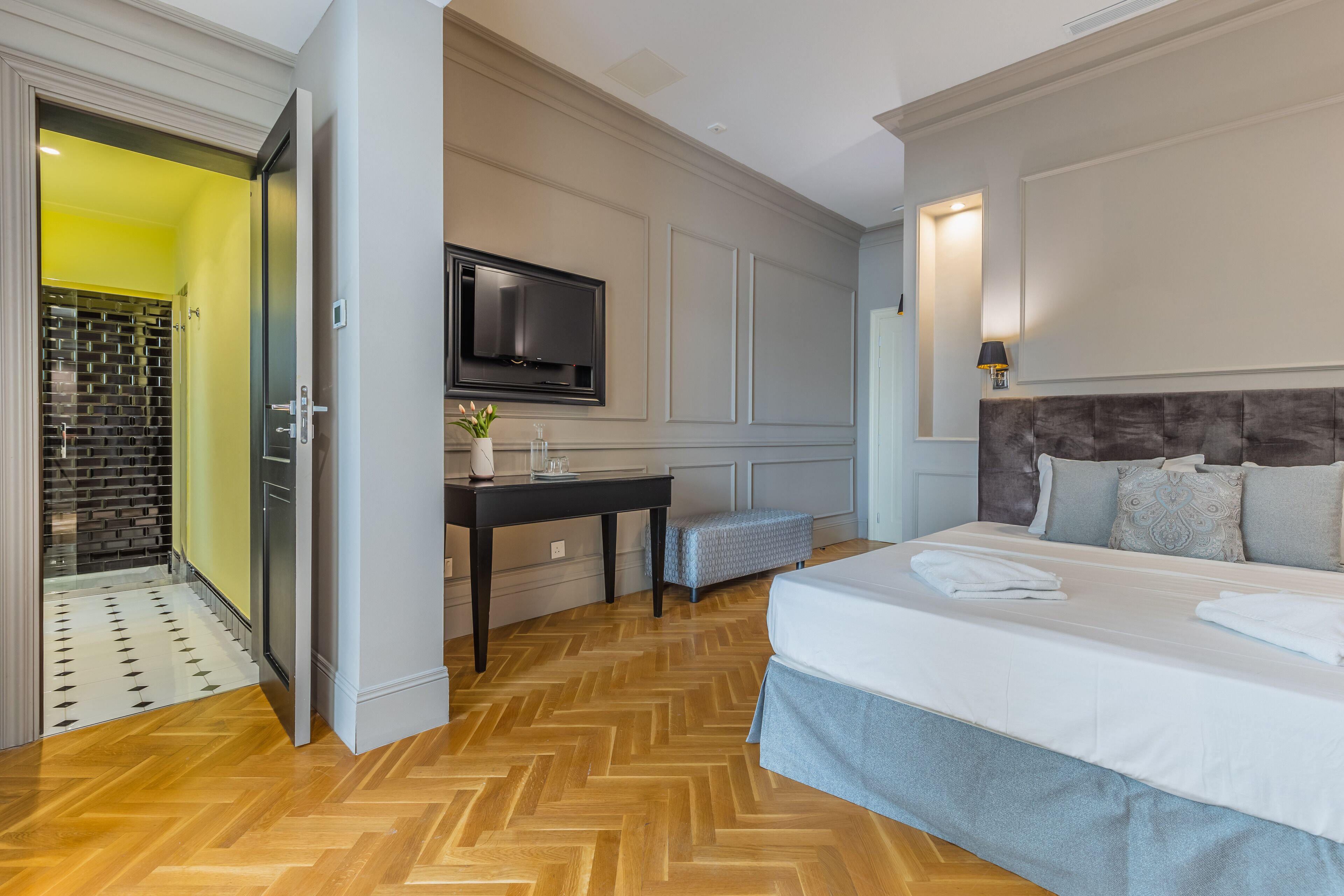 A hotel room with a large bed, a television mounted on the wall, a desk with a chair, and a bathroom with a yellow wall and a patterned floor.